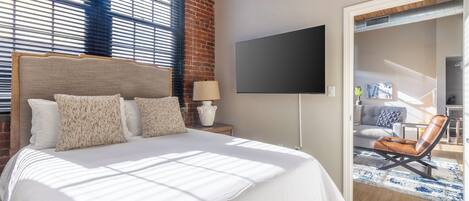 A bedroom with a large bed, a flat-screen TV, and a view of a dining area through an open door.