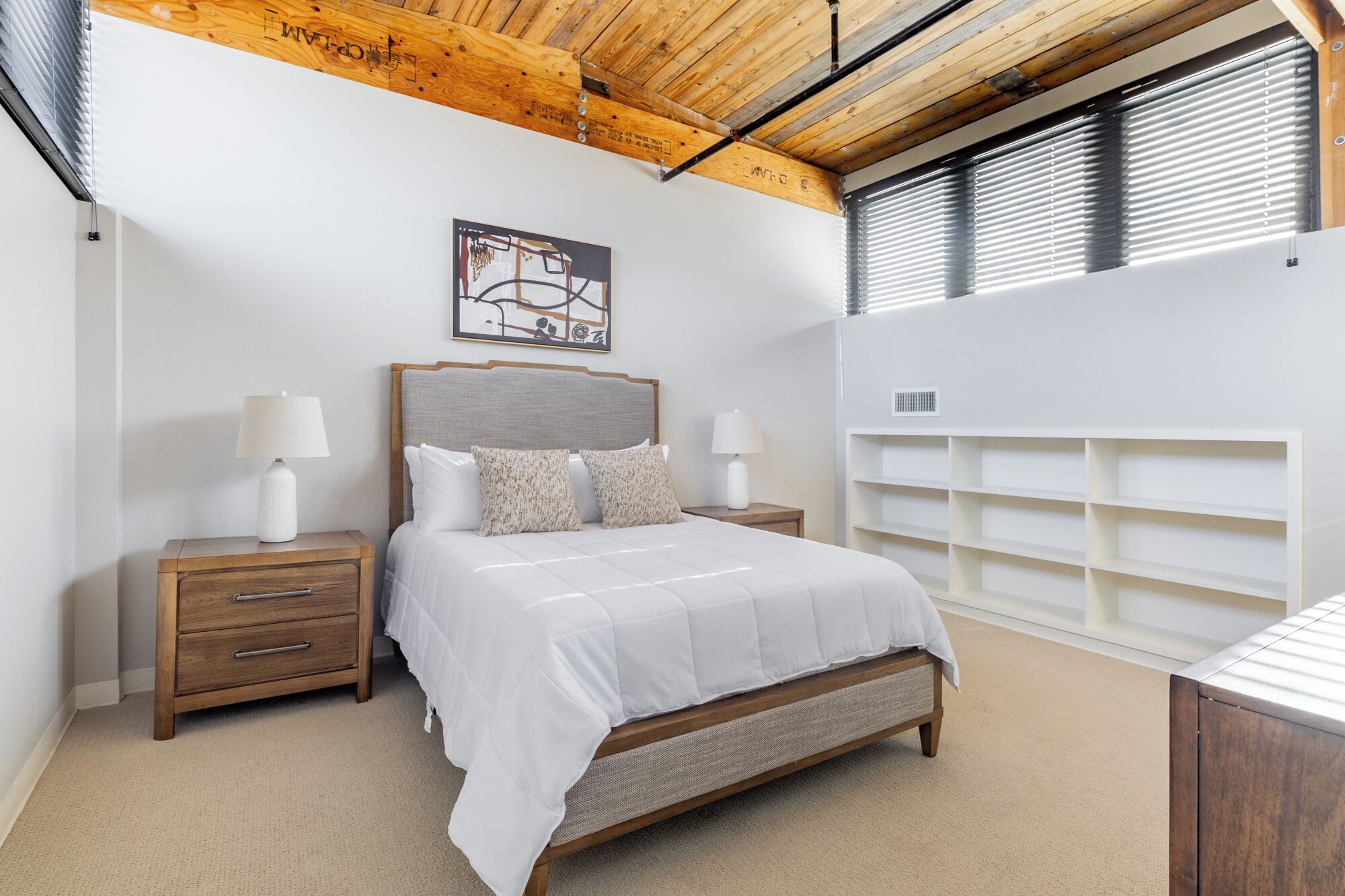 Une chambre à coucher comprenant un lit, des tables de chevet, une tête de lit, une étagère fixée au mur et un plafond avec des poutres apparentes.