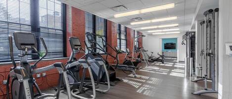 A gym with treadmills, ellipticals, and weight machines, large windows with blinds, and a clean, modern design.