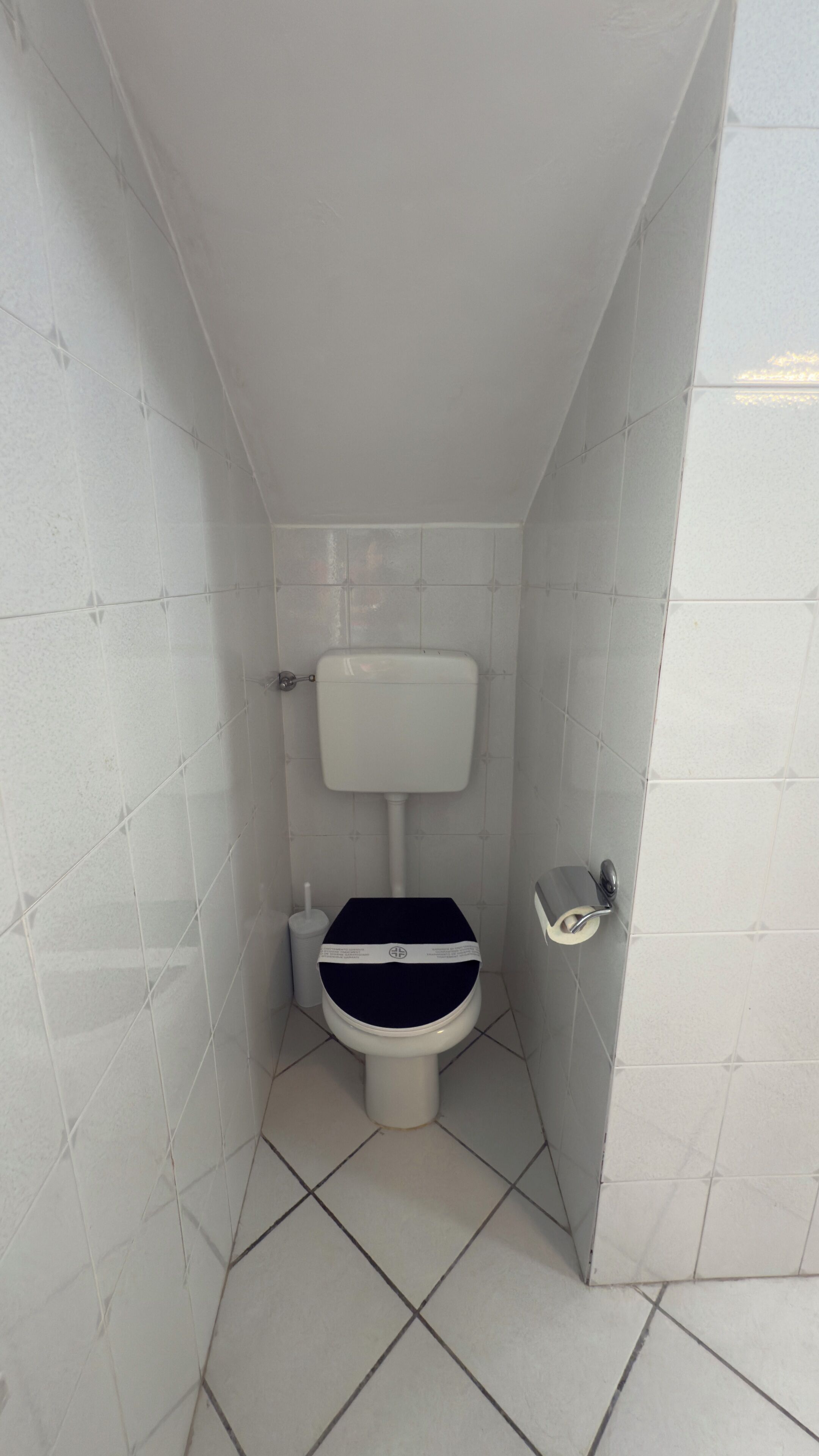 A small, enclosed bathroom with white tiled walls and floor, a white toilet, and a hand sanitizer dispenser.