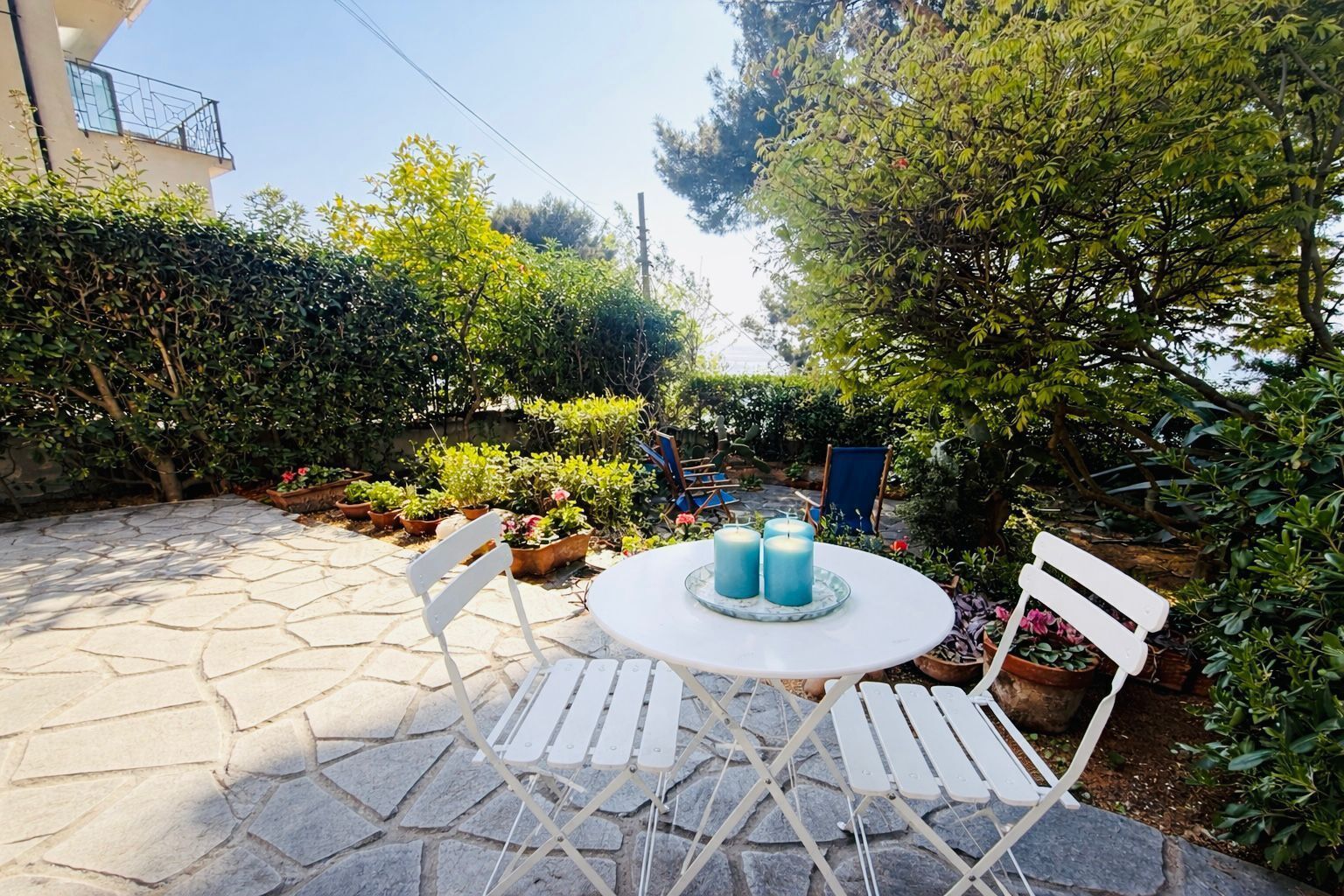 An outdoor patio with stone flooring, white chairs, a round table with candles, and potted plants.