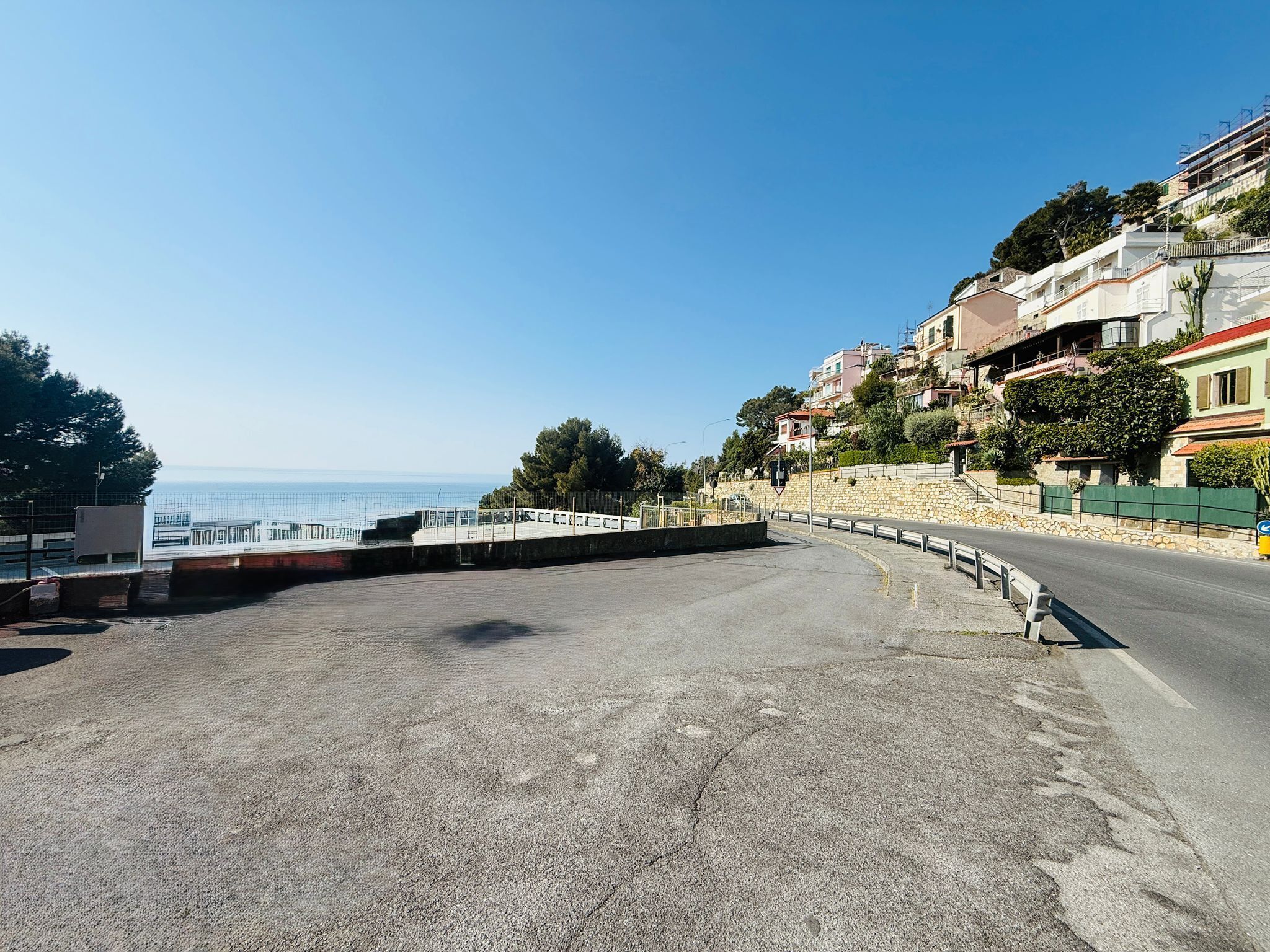 Eine Küstenstraße mit einer Steinmauer, blauem Himmel und Gebäuden am Hang.