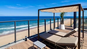 A balcony with a view of the beach and ocean, featuring lounge chairs and a potted plant. - Enjoy Salvador with the Charm of Armação (Salvador)