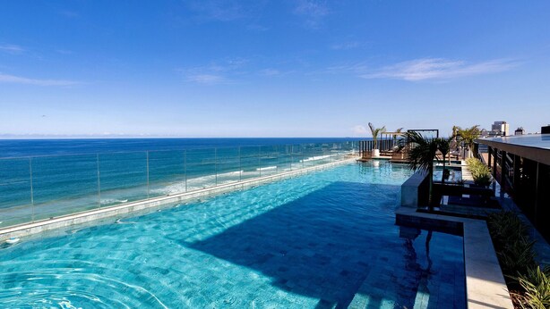 A swimming pool with a view of the ocean, a sun deck, and a lounge area with chairs and a palm tree. - Enjoy Salvador with the Charm of Armação (Salvador)