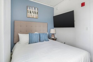 Ein Hotelzimmer mit einem Bett, einem Kopfteil, einem an der Wand montierten Fernseher, einer Nachttischlampe und einem gerahmten Bild an der Wand.