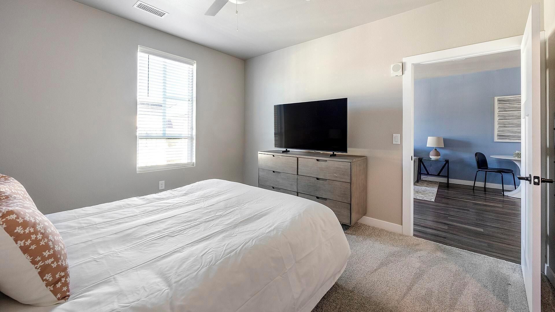 A bedroom with a bed, a TV on a dresser, a desk with a chair, and a door leading to another room.