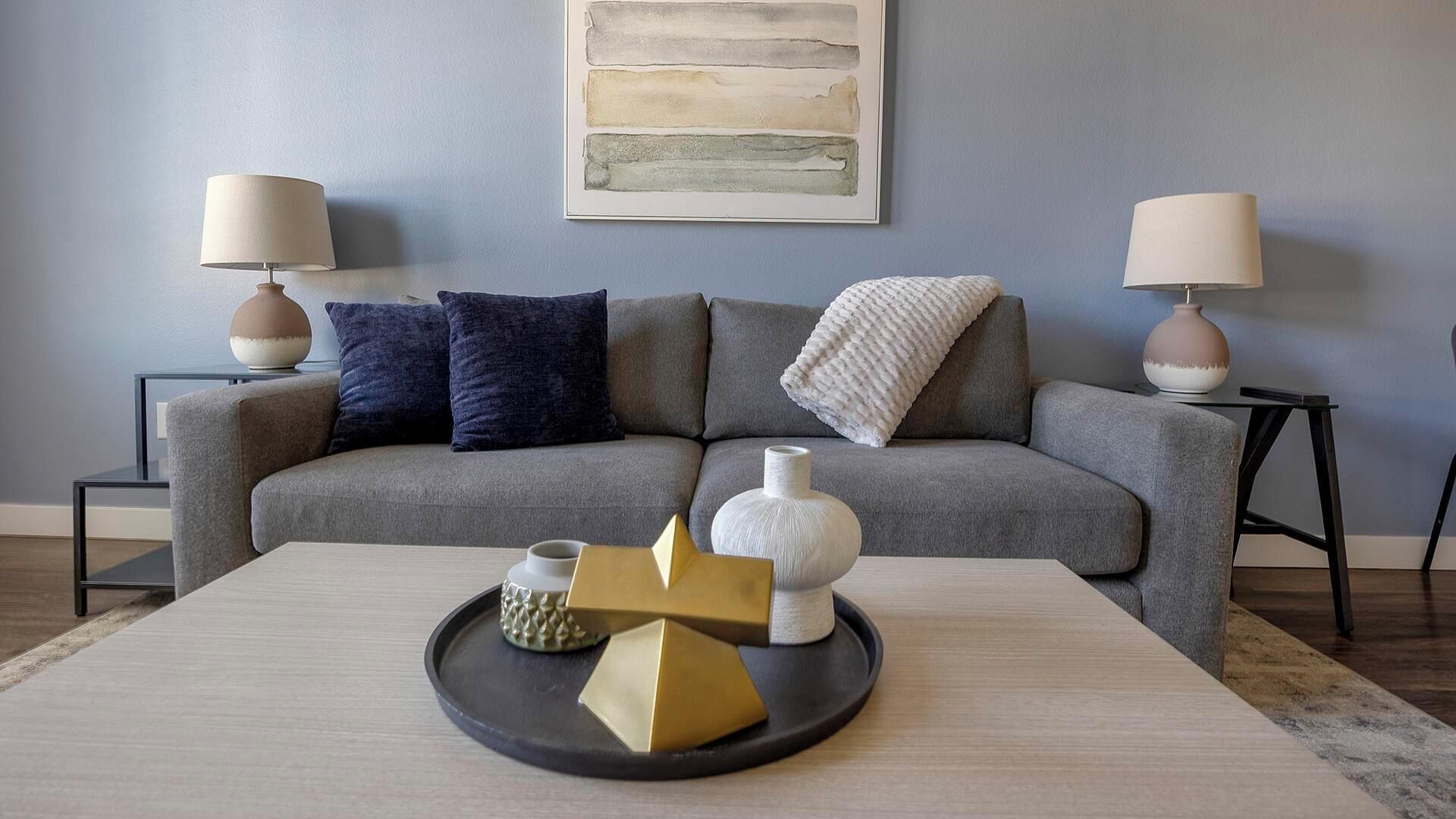 A modern living room with a grey sofa, two side tables with lamps, a coffee table with decorative items, and a wall art piece.