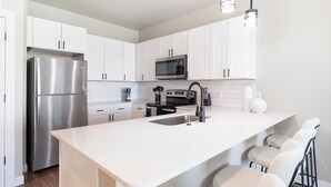 A modern kitchen with white cabinets, stainless steel appliances, a central island with bar stools, and pendant lighting. - Landing | Incredible 2BD, Clubhouse (Goodyear)