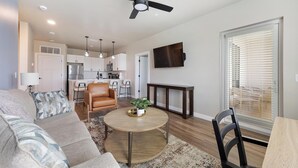 A modern living room with a sofa, a coffee table with a plant, a dining area with a table and chairs, a kitchen with white cabinets, and a ceiling fan. - Landing | Incredible 2BD, Clubhouse (Goodyear)