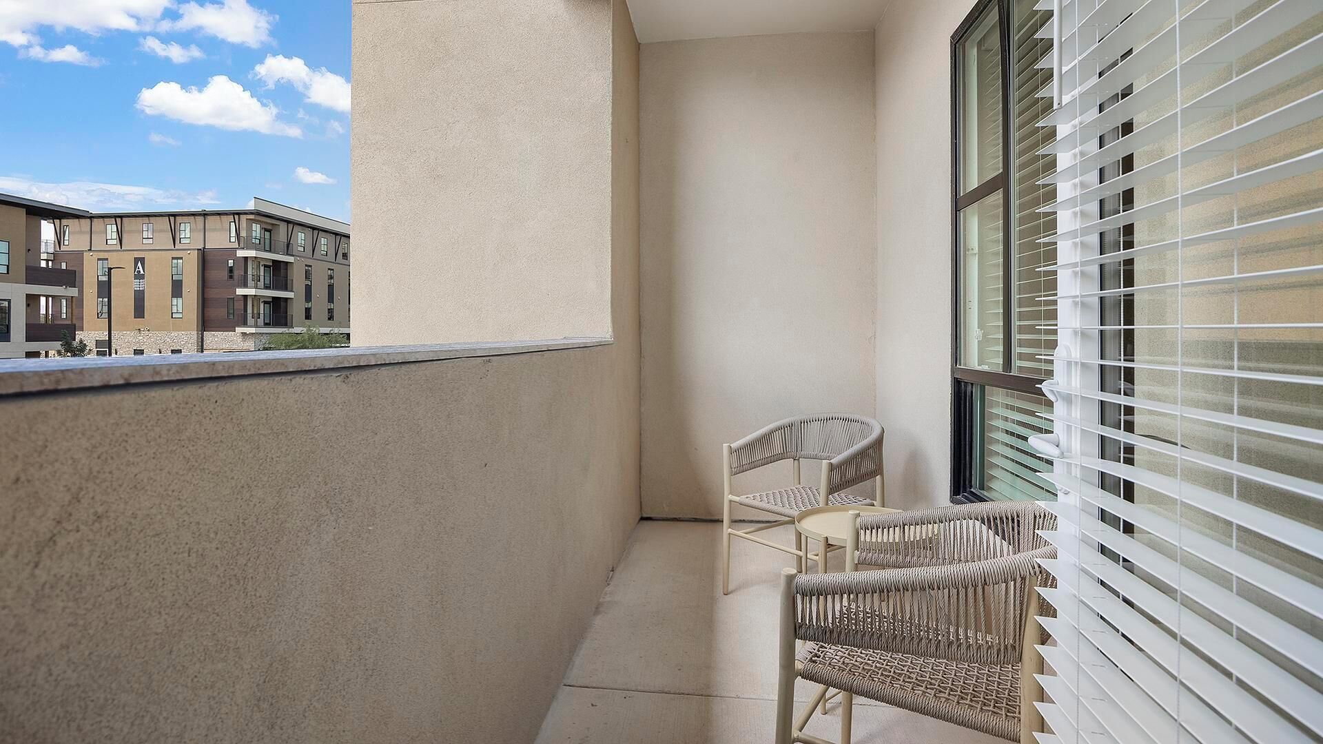 Un balcon avec deux fauteuils en osier, une fenêtre avec des stores et une vue sur les immeubles voisins.
