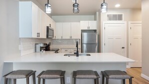A modern kitchen with white cabinets, a stainless steel refrigerator, a central island with bar stools, and pendant lighting. - Landing | Stylish 2BD, Clubhouse (Goodyear)
