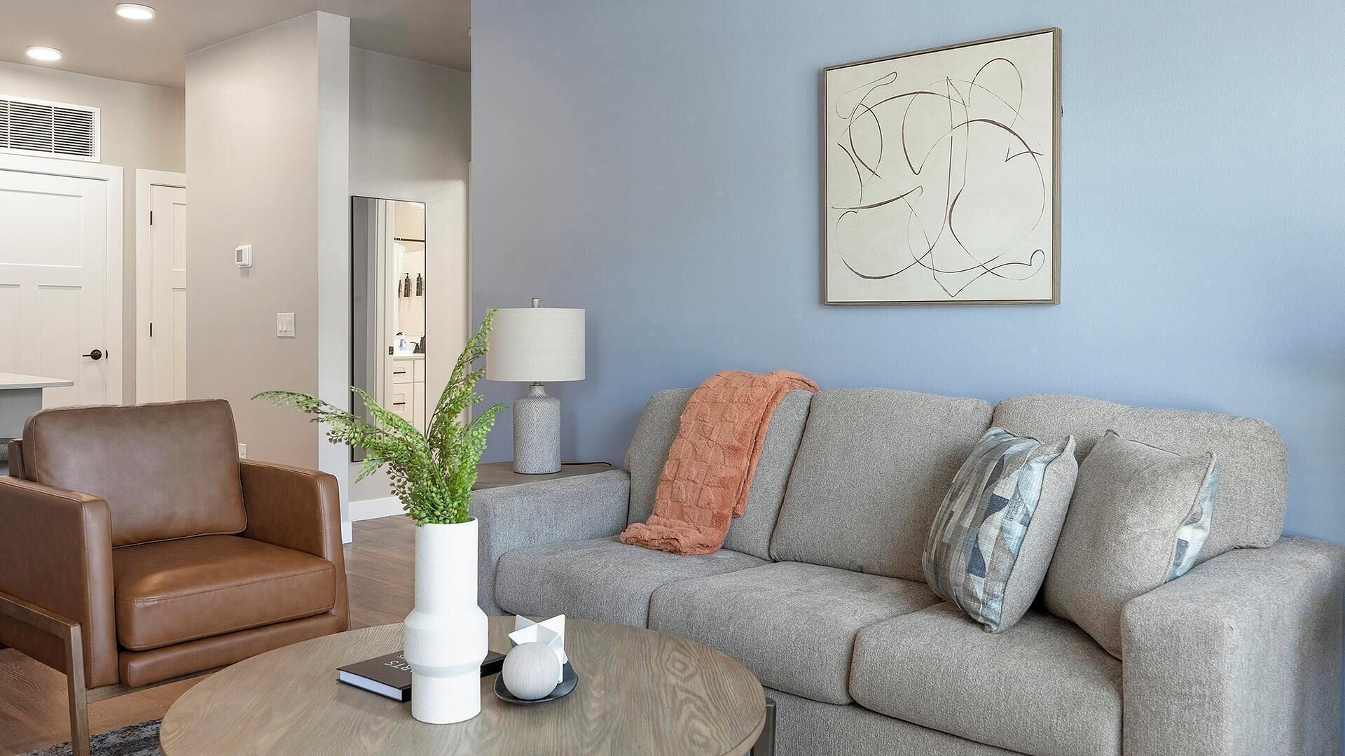 A living room with a grey sofa, a brown leather chair, a round wooden coffee table, and a white vase with a plant.