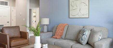 A living room with a grey sofa, a brown leather chair, a round wooden coffee table, and a white vase with a plant.