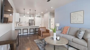 A modern living room with a grey sofa, a coffee table, and a brown leather chair. There is a kitchen area with white cabinets and stainless steel appliances. - Landing | Stylish 2BD, Clubhouse (Goodyear)
