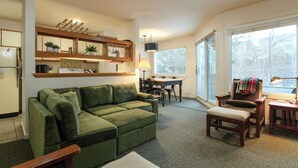 A cozy living room with a green sofa, a dining table, and a chair. There is a kitchen area with a refrigerator and a shelf with plants. - Sugarbush Escape  Fireplace and Shuttle Access (Warren)