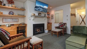A living room with a fireplace, wooden furniture, a television mounted above the fireplace, and a green couch. - Sugarbush Escape  Fireplace and Shuttle Access (Warren)