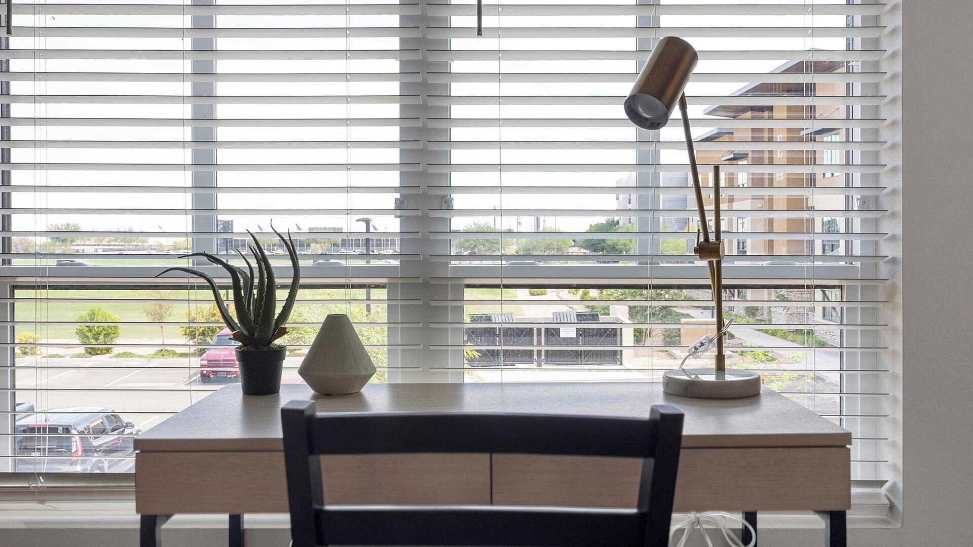 A desk with a chair, a lamp, and two potted plants in front of a window with horizontal blinds.