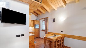 Une cuisine avec des meubles en bois, une table à manger avec des chaises, un téléviseur fixé au mur et une fenêtre avec des rideaux.