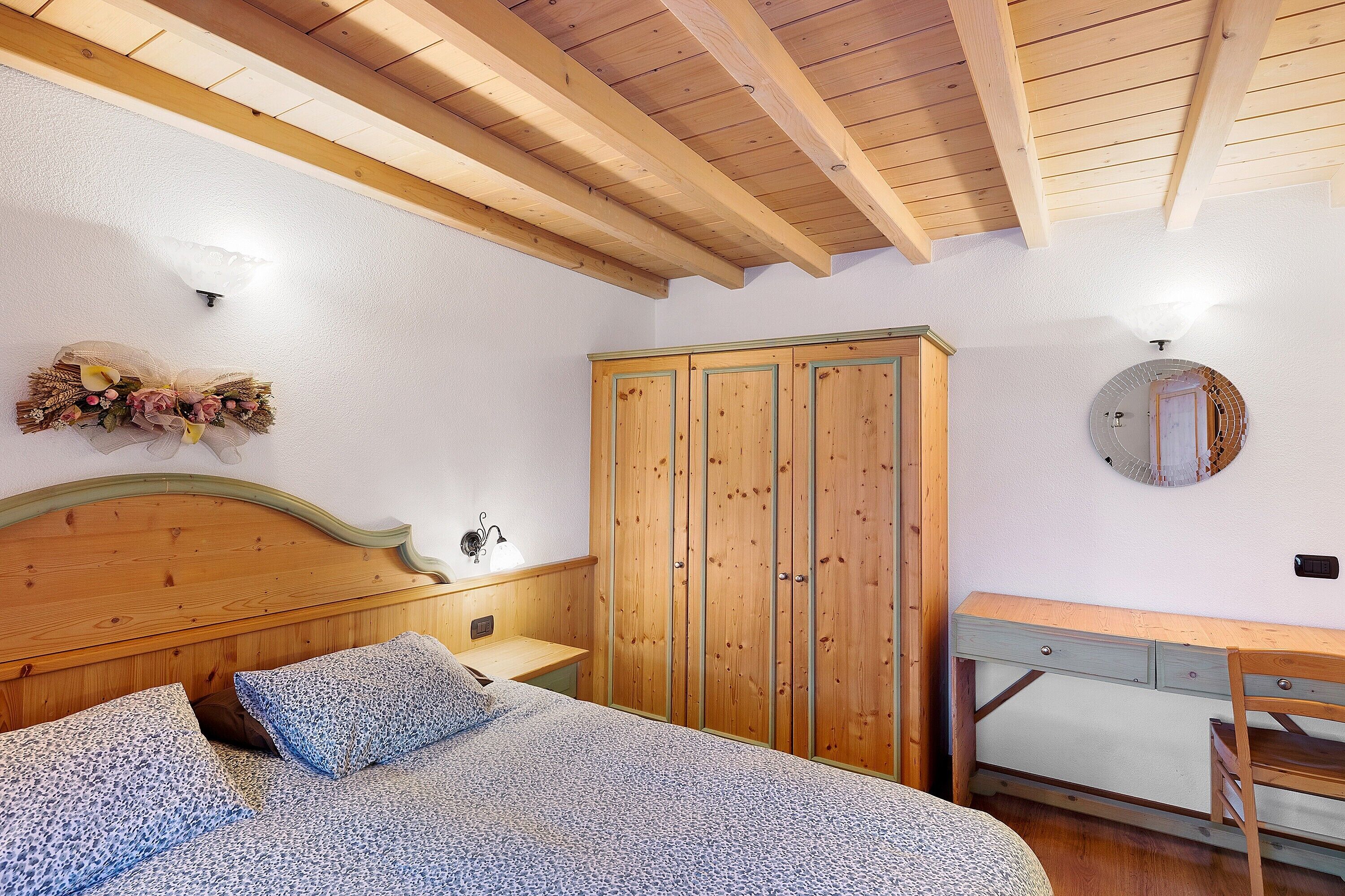 A bedroom with a wooden bed, a wooden wardrobe, a desk, and a mirror on the wall.