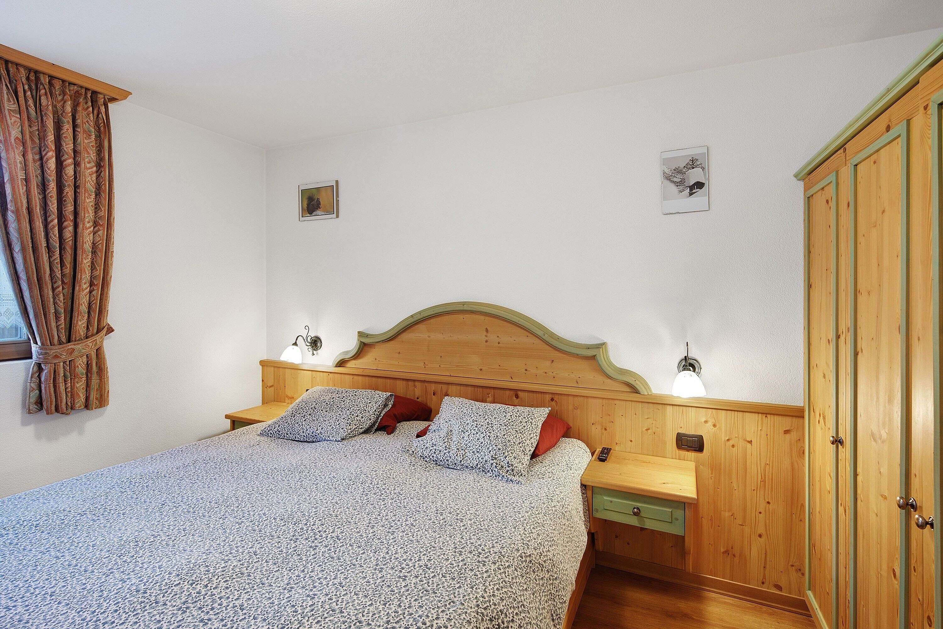 Une chambre à coucher avec une tête de lit en bois, un lit recouvert d’une literie à motifs, deux lampes de chevet, une table de chevet et une armoire.