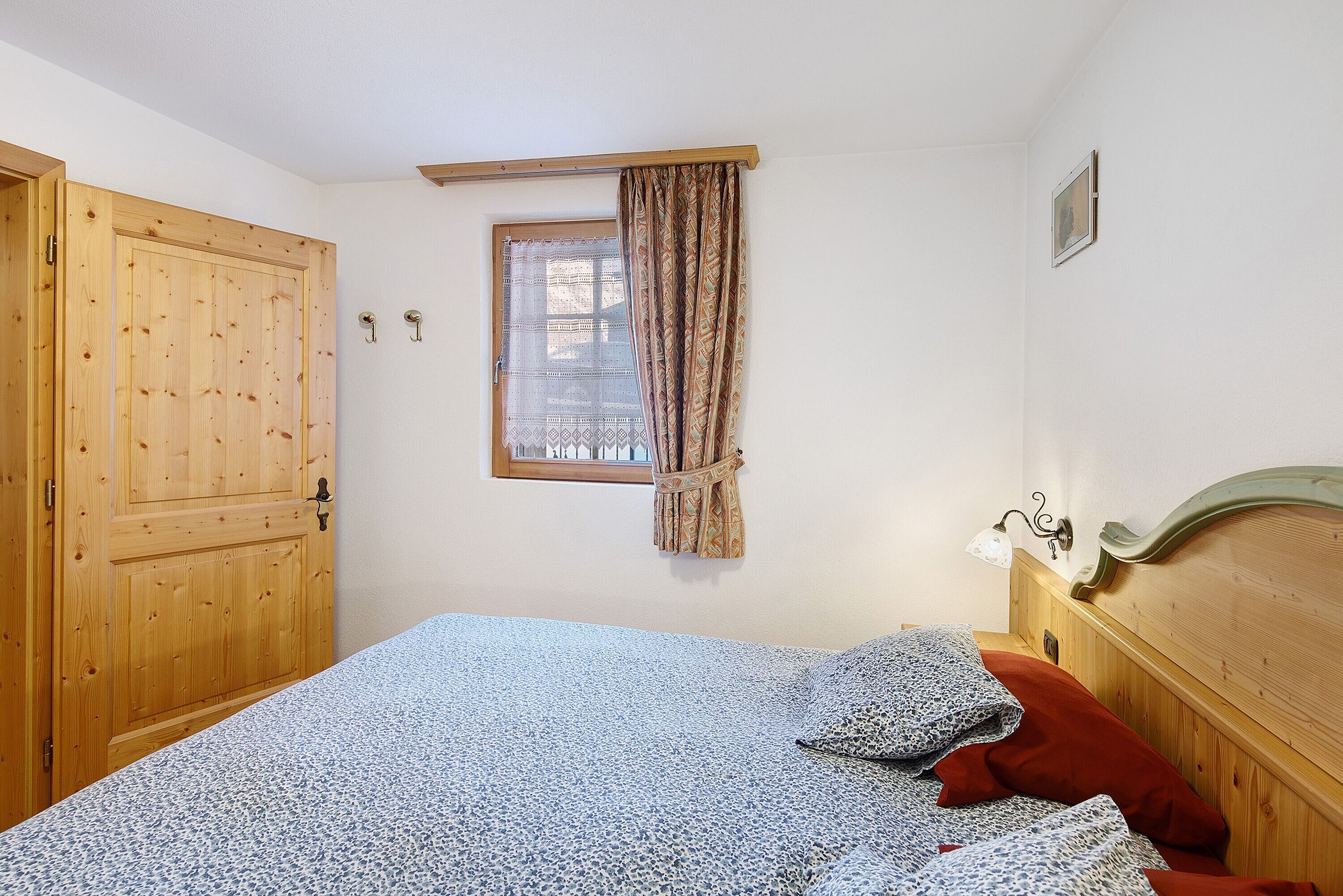 A bedroom with a wooden door, a window with patterned curtains, a bed with patterned bedding, and a headboard with a light fixture.
