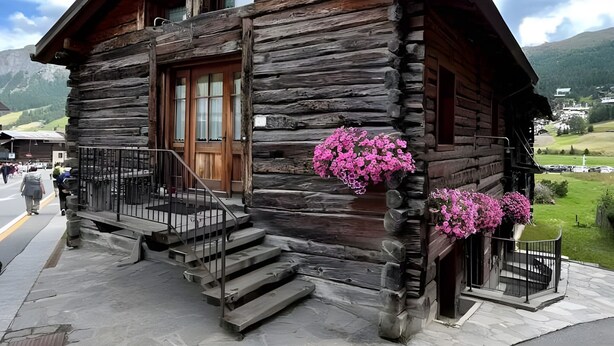 A rustic log cabin with a wooden door, a balcony with pink flowers, and a stone-paved entrance. - Apartment 'Coccinella 2' with Mountain View (Livigno)