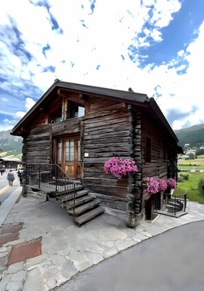 Ein rustikales Blockhaus mit Holztür, einem Balkon mit rosa Blumen und einem eingepflasterten Zugang.