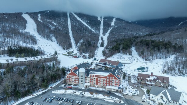 A large, red-brick hotel with multiple wings and a central tower, surrounded by snowy mountain terrain and a parking lot. - Village Edge – Steps to the slopes at Sugarbush! (Warren)