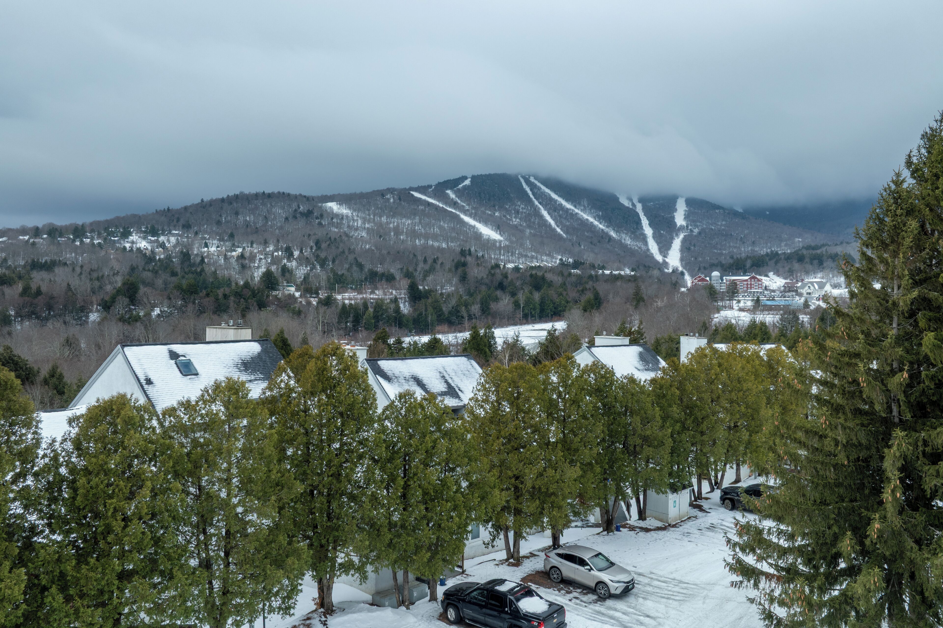 A snowy landscape with a mountain in the background, a parking area with cars, and trees in the foreground.