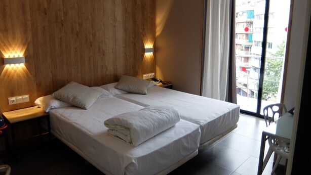 A hotel room with a large bed, wooden panel walls, wall-mounted lights, a bedside table, and a view of the city through a large window. - HOTEL MEDICIS (Barcelona)