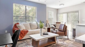 Ein modernes Wohnzimmer mit einer Couch, einem Couchtisch und einem Sessel. Große Fenster mit Jalousien, eine Lampe und ein kleiner Tisch sind ebenfalls vorhanden.