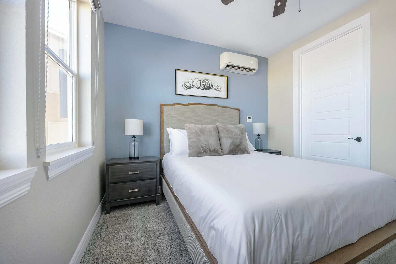 A bedroom with a bed, two bedside tables, a nightstand, a wall-mounted air conditioner, and a door.