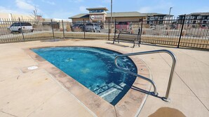 Una piscina con barandilla metálica, rodeada por una valla, y un edificio con la leyenda 'Apartment' al fondo.