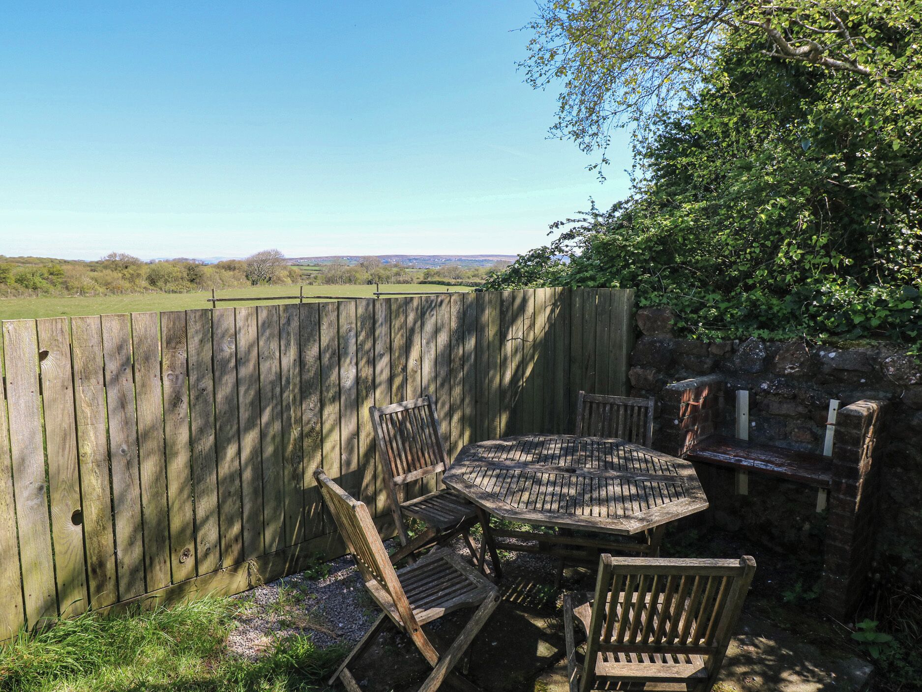 A wooden table and chairs in a garden with a wooden fence and a stone wall.