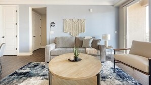 Un salon avec un canapé gris, une table basse ronde en bois, une plante en pot et une décoration murale en macramé.