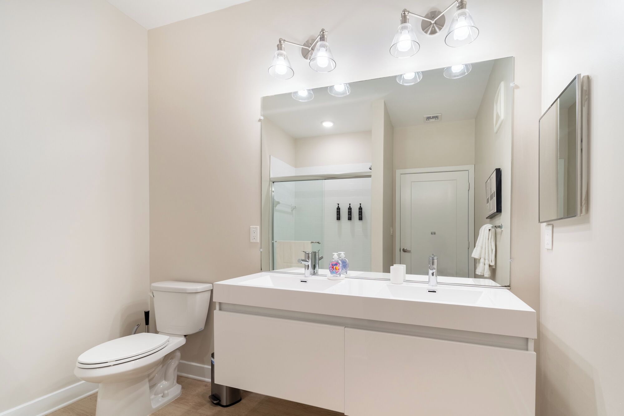 Casa de banho moderna com lavatório branco, espelho e sanita. Três luzes acima do espelho e um porta-toalhas na parede.