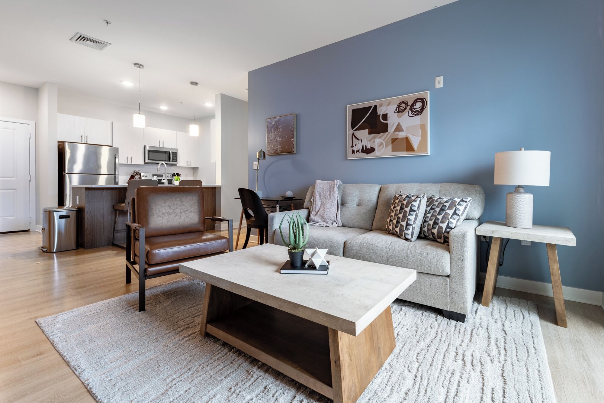 A modern living room with a sofa, coffee table, and chairs. There is a kitchen area with a refrigerator and microwave in the background.