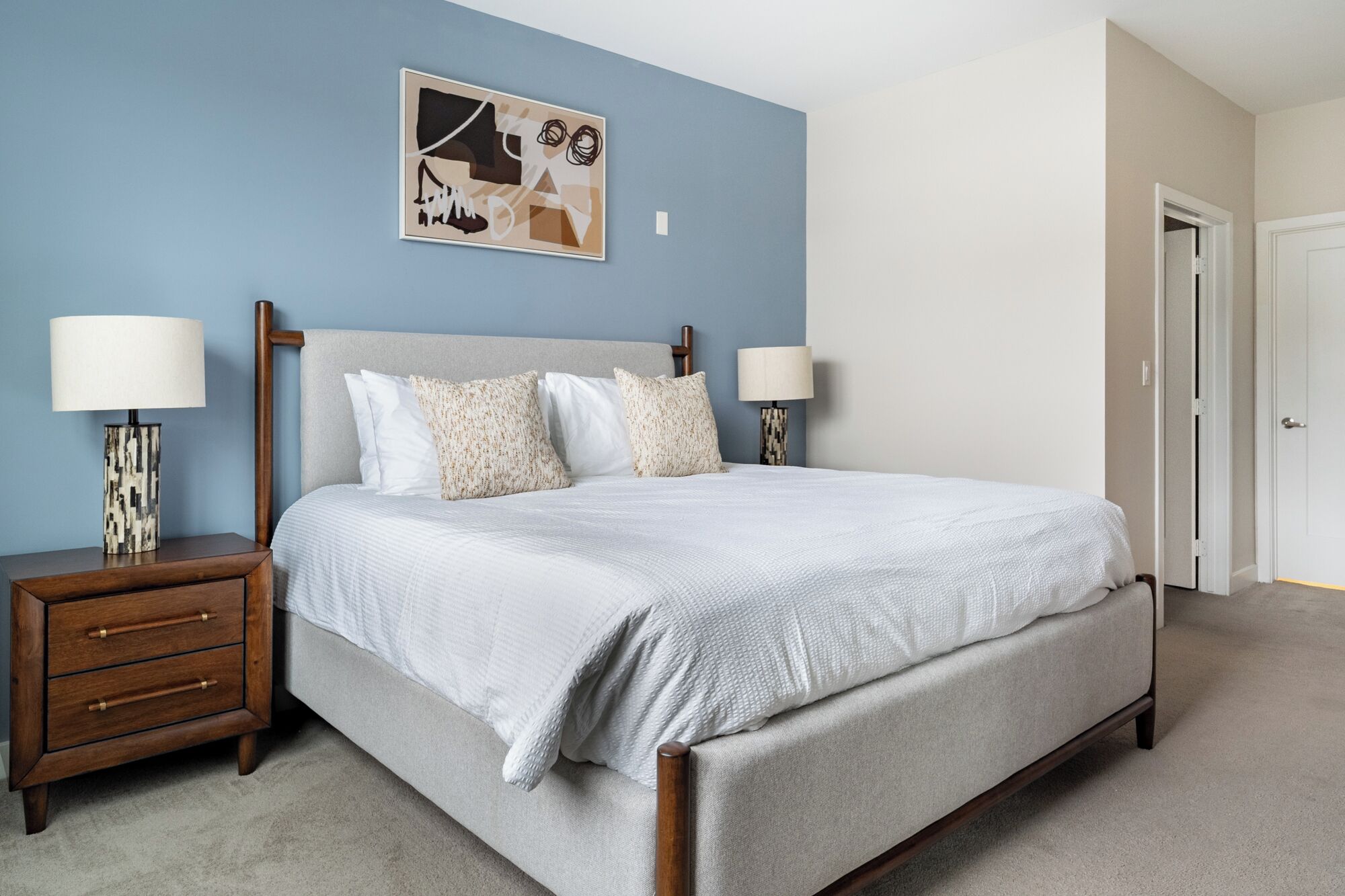 A bedroom with a bed, two bedside tables, a nightstand, a door, and a wall with a framed picture.