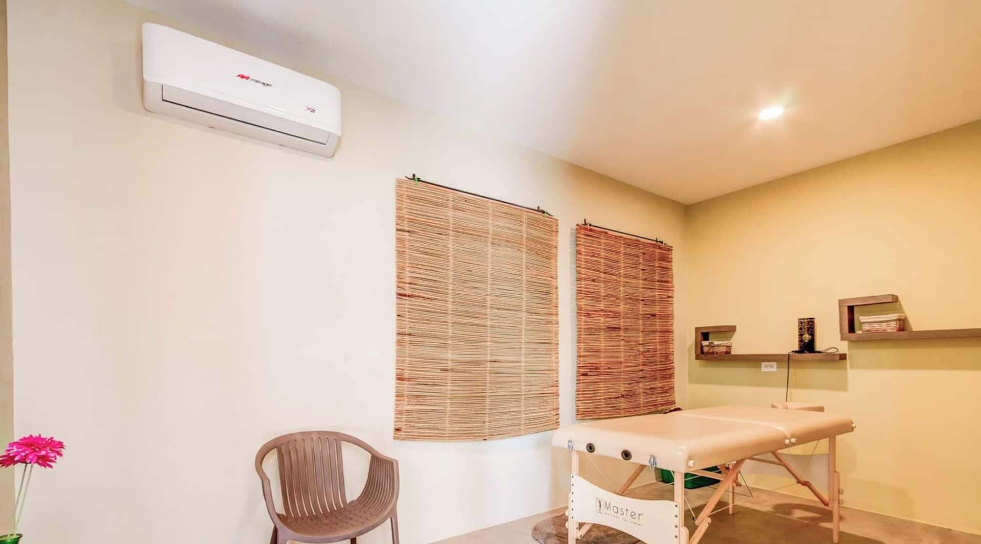 Une pièce comprenant une table de massage, une chaise, un climatiseur fixé au mur et deux stores en bambou.