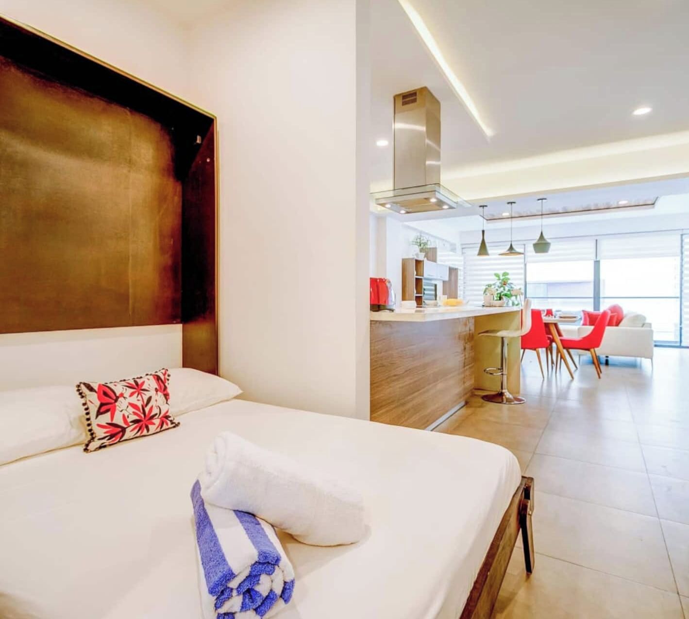 A modern hotel room with a large bed, a kitchenette, and a dining area with red chairs.