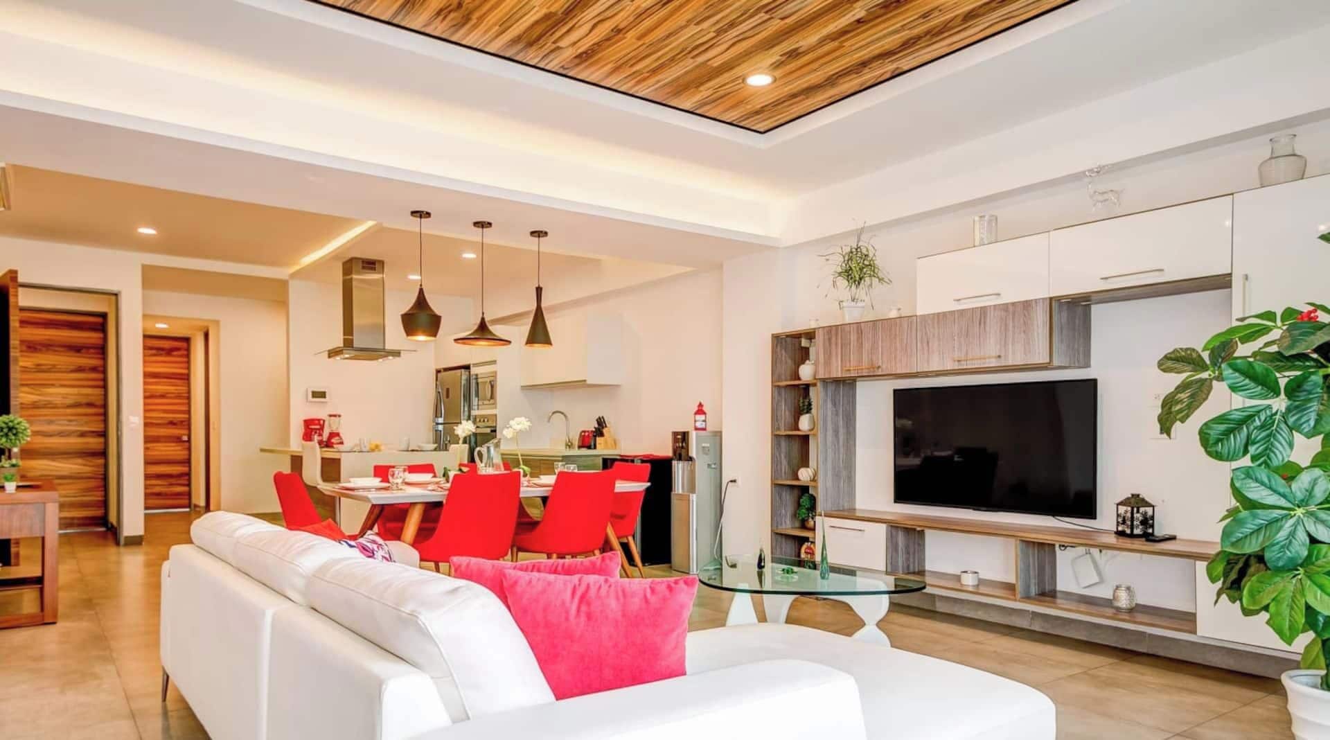 Ein modernes Wohnzimmer mit einem weißen Sofa, roten Sesseln, einem Glastisch und einem großen Flachbildfernseher, der an einem Holzschrank befestigt ist.