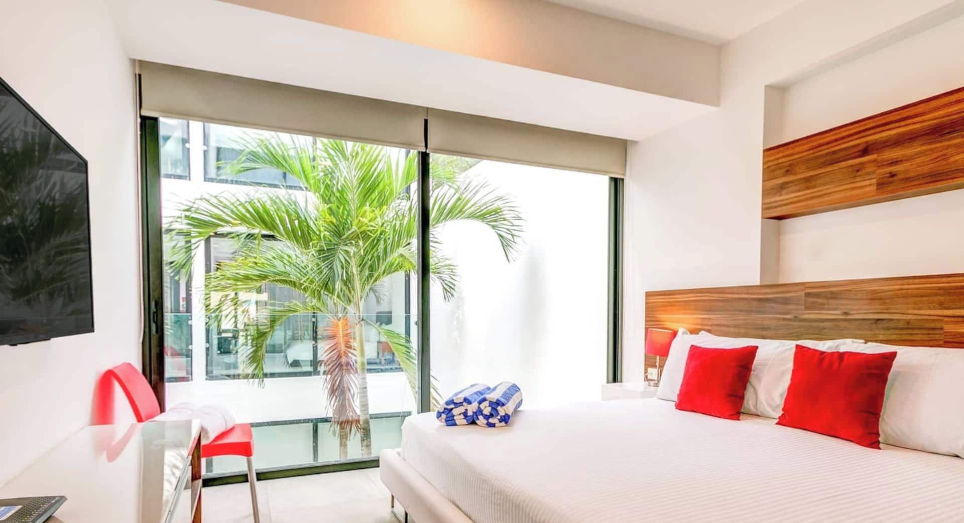 Een moderne hotelkamer met een groot bed, een bureau, een stoel en een groot raam met uitzicht op palmbomen.