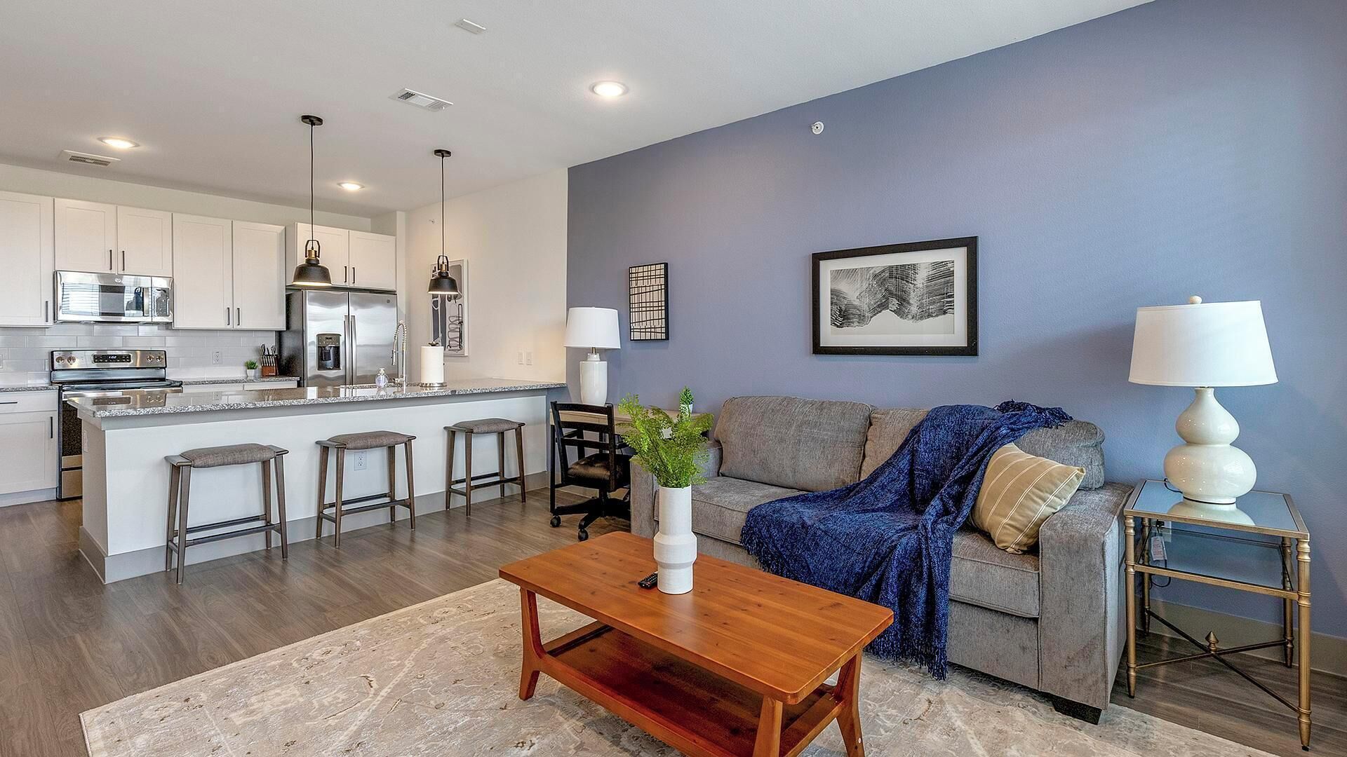 A modern living room with a grey sofa, a wooden coffee table, and a kitchen area with white cabinets and stainless steel appliances.