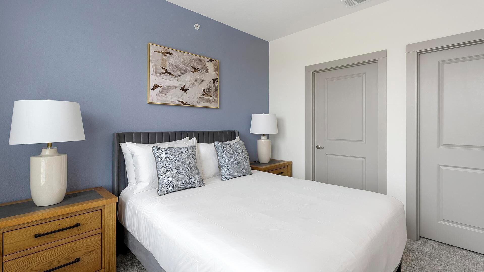 A neatly arranged bedroom with a bed, two bedside tables with lamps, a wall-mounted painting, and a door.