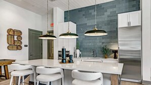 Una cocina moderna con una isla grande, taburetes blancos tipo bar, luces colgantes y un refrigerador de acero inoxidable.