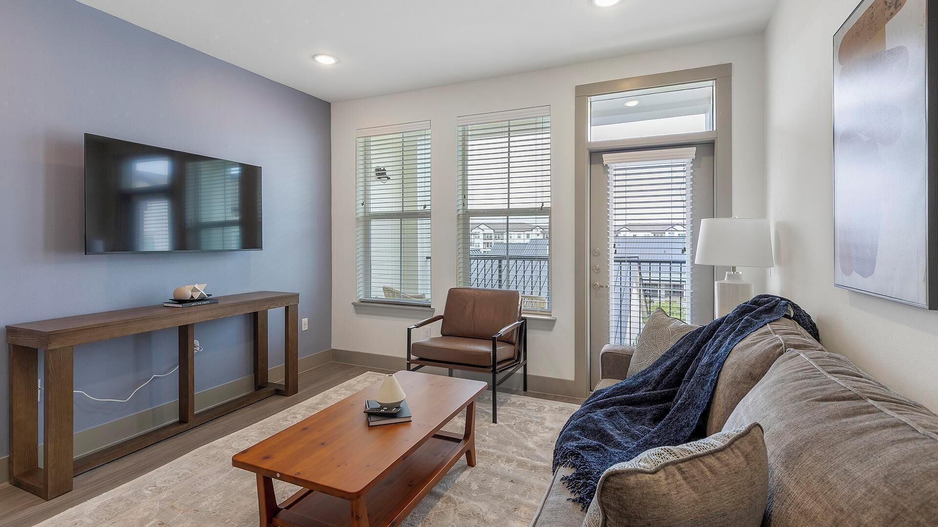 A modern living room with a flat-screen TV, a wooden console table, a leather armchair, a sofa, and large windows with blinds.
