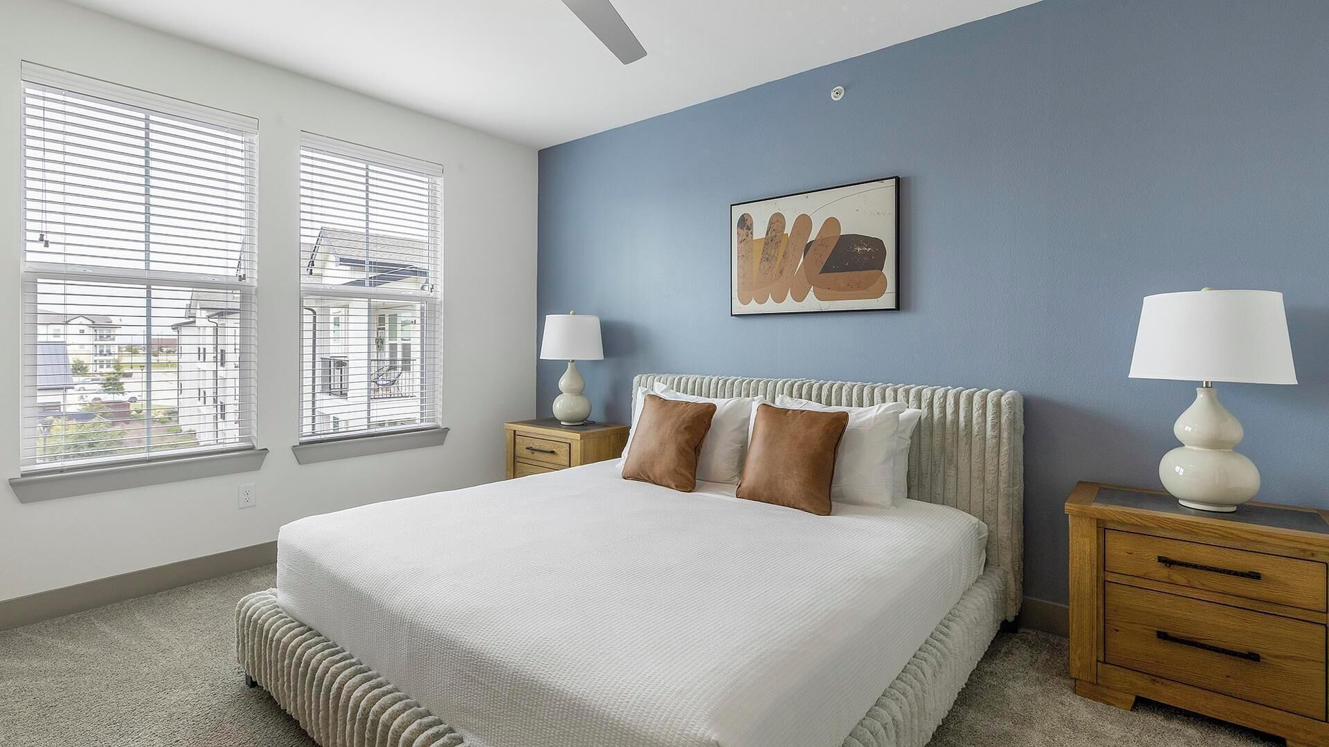 Une chambre à coucher avec un grand lit, deux tables de chevet, une fenêtre avec des stores, une lampe fixée au mur et un tableau encadré.