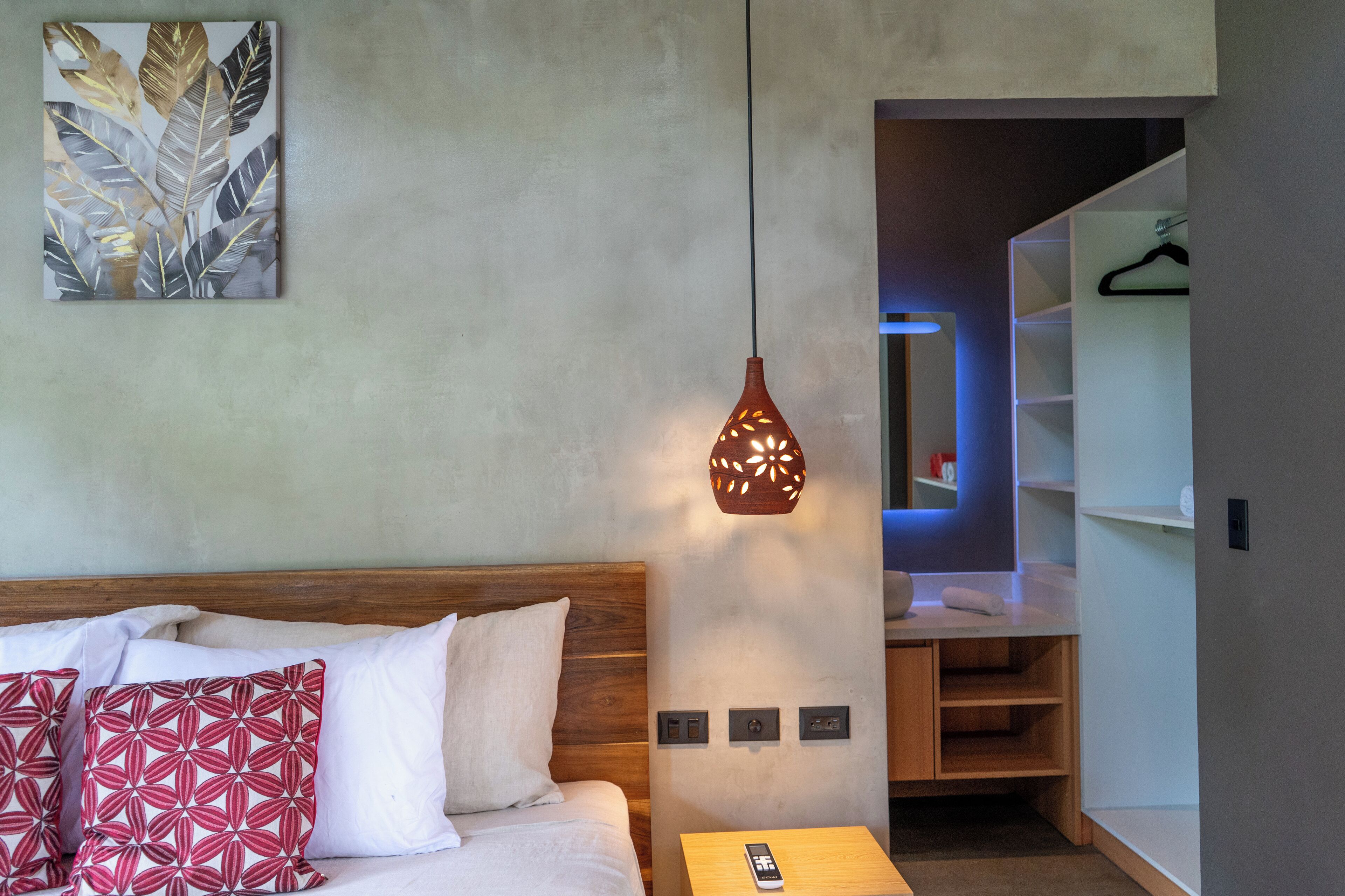 A modern hotel room with a wooden headboard, a bed with white and red patterned pillows, a nightstand with a remote control, and a wall-mounted artwork.
