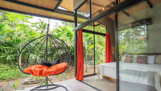 Ein Schlafzimmer mit einem großen Glasfenster, einem Hängesessel und Blick auf üppiges Grün.