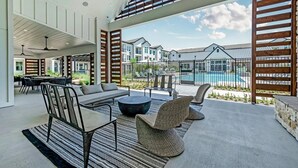 Amplo espaço ao ar livre com mesa, cadeiras e um sofá. Dá para ver uma piscina e outros edifícios ao fundo.